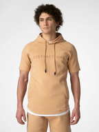 A man wearing a tan short-sleeve pullover hoodie with 'DISTINCT ONE' printed across the chest, paired with matching tan pants, against a light gray studio background.