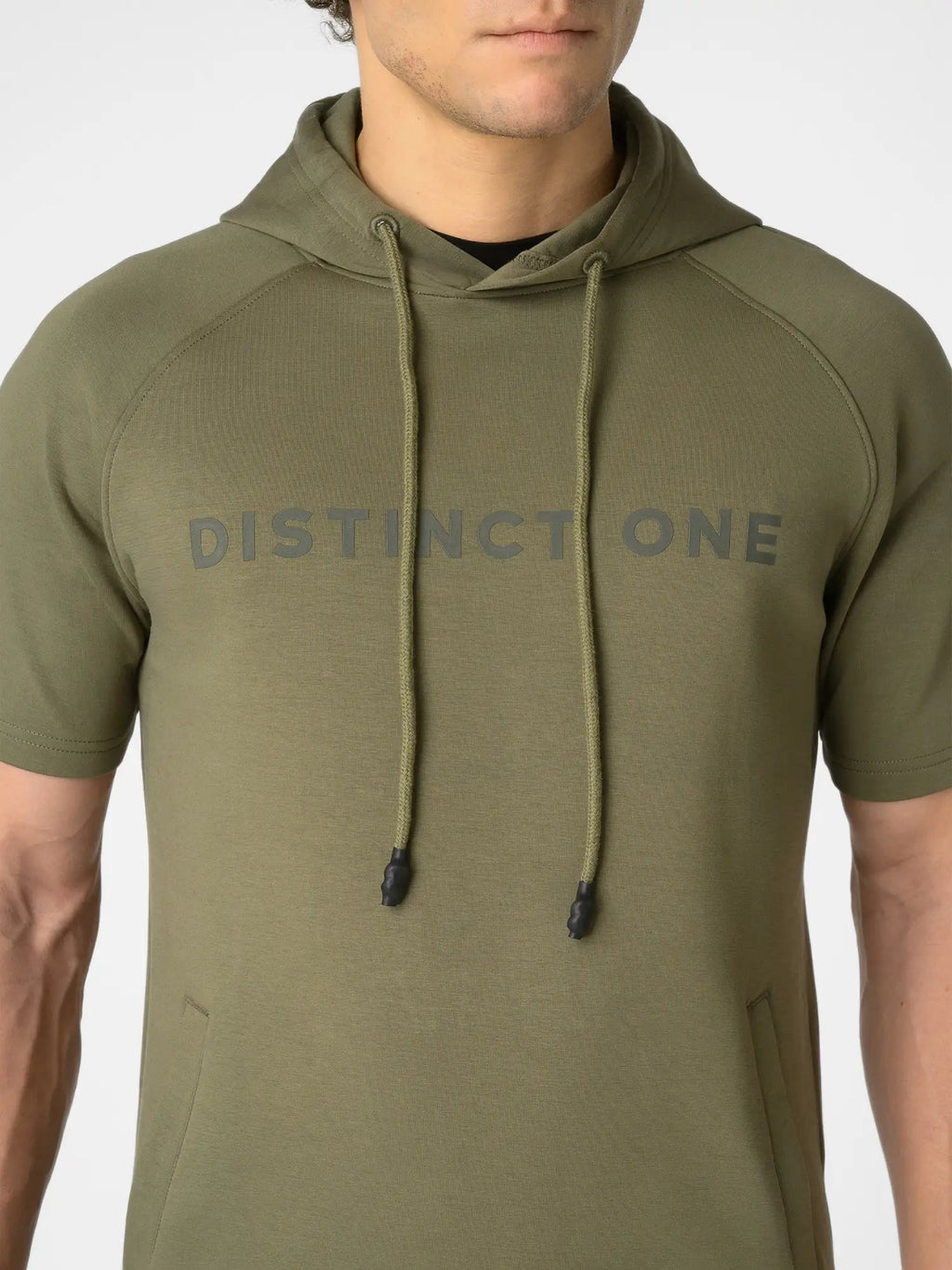 A man wearing an olive green short-sleeve hoodie with 'DISTINCT ONE' printed in gray across the chest, shown from the neck down against a light gray background.