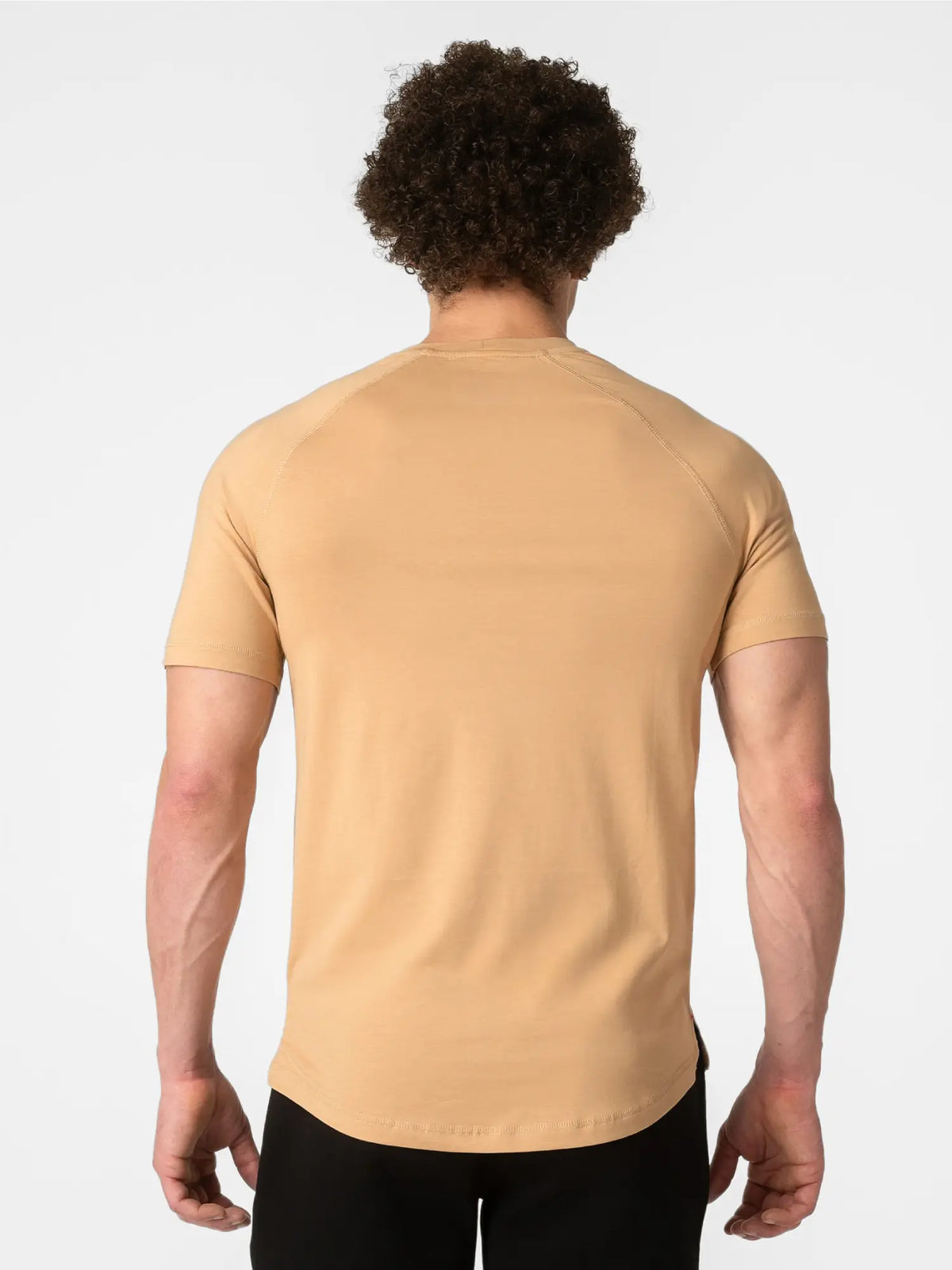 A man viewed from behind wearing a short-sleeve crew-neck t-shirt in a warm tan/beige color, paired with black pants, against a light gray studio background.