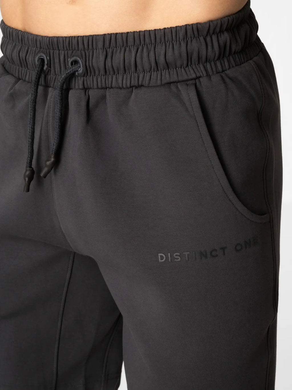 Close-up of a person wearing dark charcoal gray sweatpants with an elastic drawstring waistband and a subtle tonal 'DISTINCT ONE' logo on the thigh.