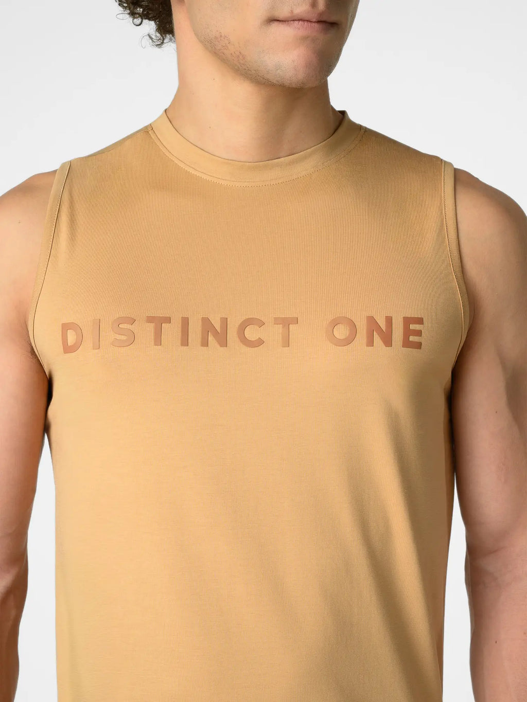 A close-up of a man wearing a tan sleeveless muscle tank top with the words 'DISTINCT ONE' printed across the chest in a matching tan color.