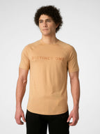 A man wearing a tan short-sleeve athletic t-shirt with 'DISTINCT ONE' printed across the chest in a darker tan tone, paired with black pants, against a light gray studio background.