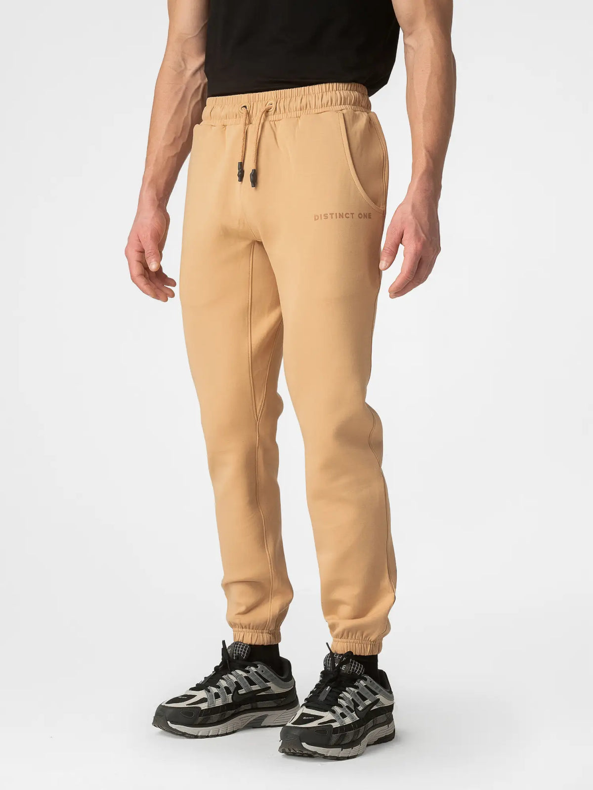 A person wearing beige jogger sweatpants with a drawstring waist, side pockets, and elastic cuffs, paired with black and gray sneakers against a light gray studio background.