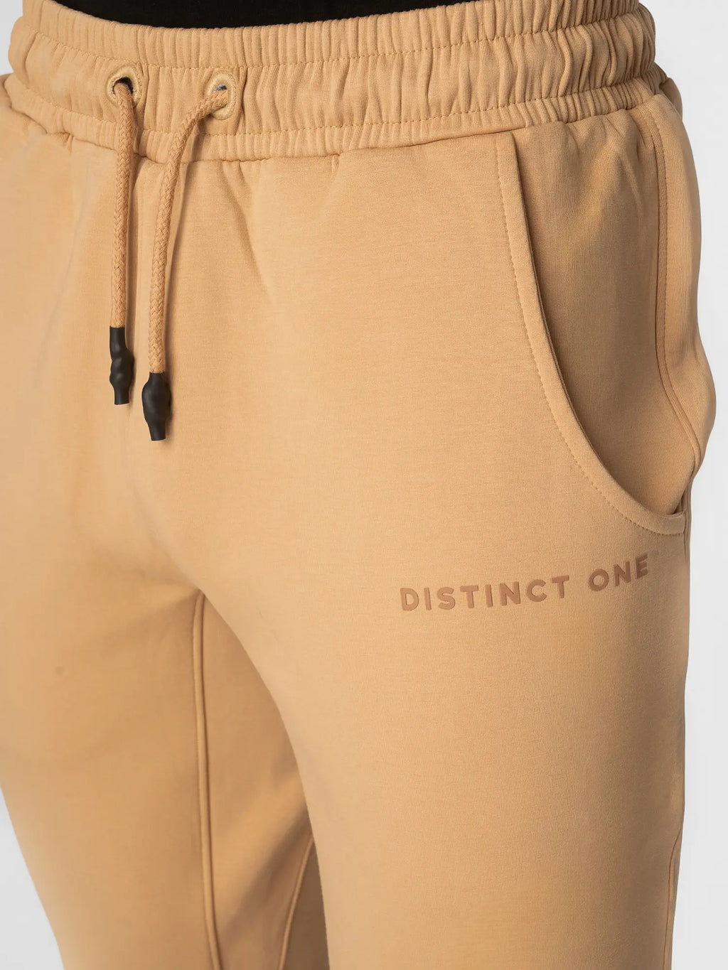 Close-up of tan sweatpants with a drawstring waist, side pocket, and 'DISTINCT ONE' printed on the thigh.