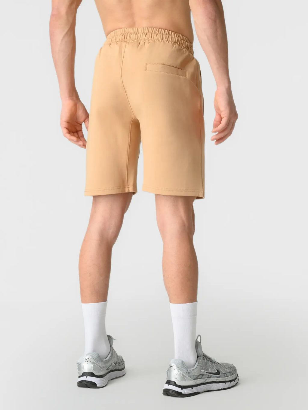 A man is shown from the back wearing light peach-beige athletic shorts, white socks, and silver running shoes against a light gray studio background.