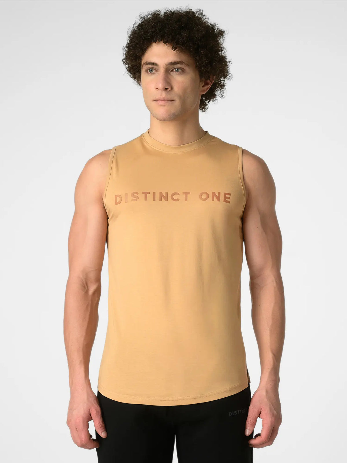 A man wearing a sleeveless tan athletic tank top with 'DISTINCT ONE' printed across the chest in a darker tan, paired with black pants, against a light gray studio background.