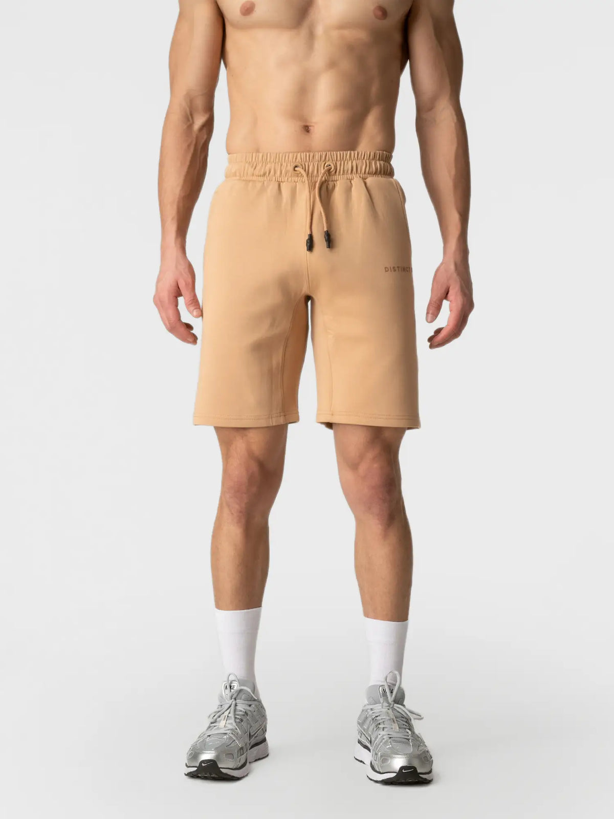 A man wearing beige athletic shorts with a drawstring waist, white socks, and gray sneakers, shown from the chest down against a light gray studio background.