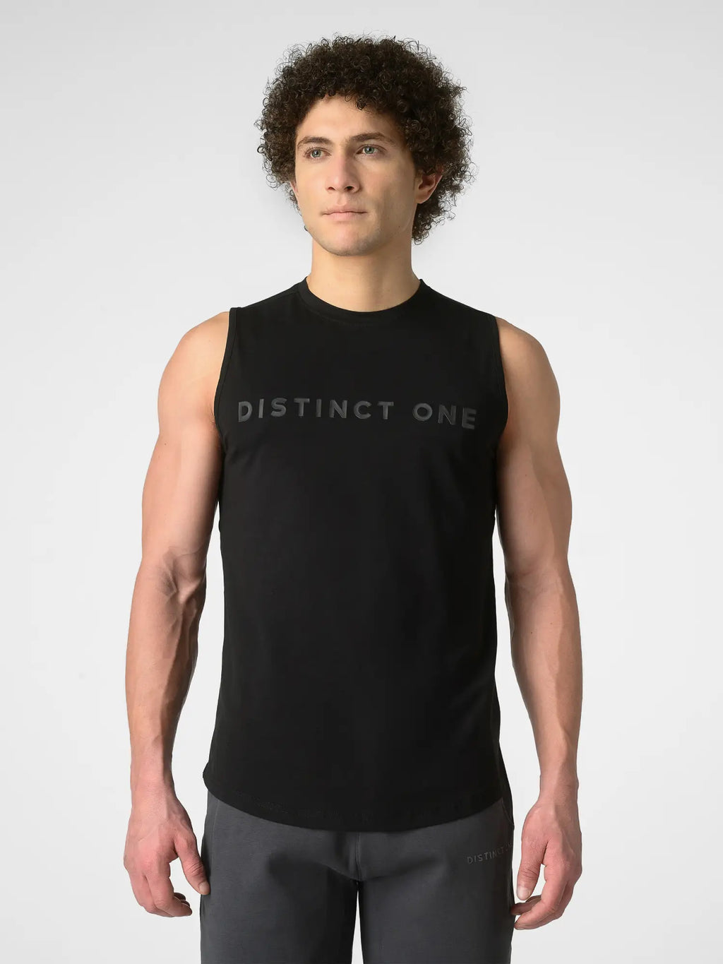 A man wearing a black sleeveless athletic tank top with 'DISTINCT ONE' printed across the chest, paired with dark gray athletic pants, against a light gray studio background.