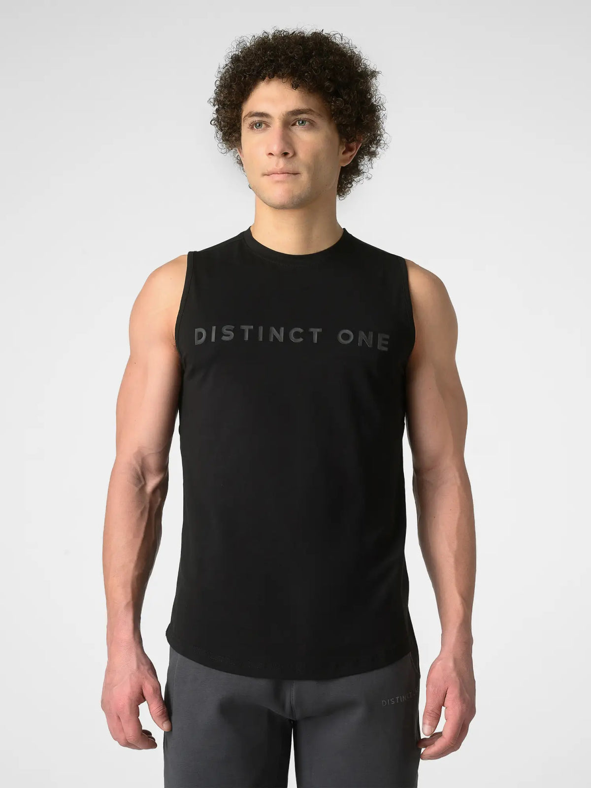 A man wearing a black sleeveless athletic tank top with 'DISTINCT ONE' printed across the chest, paired with dark gray athletic pants, against a light gray studio background.