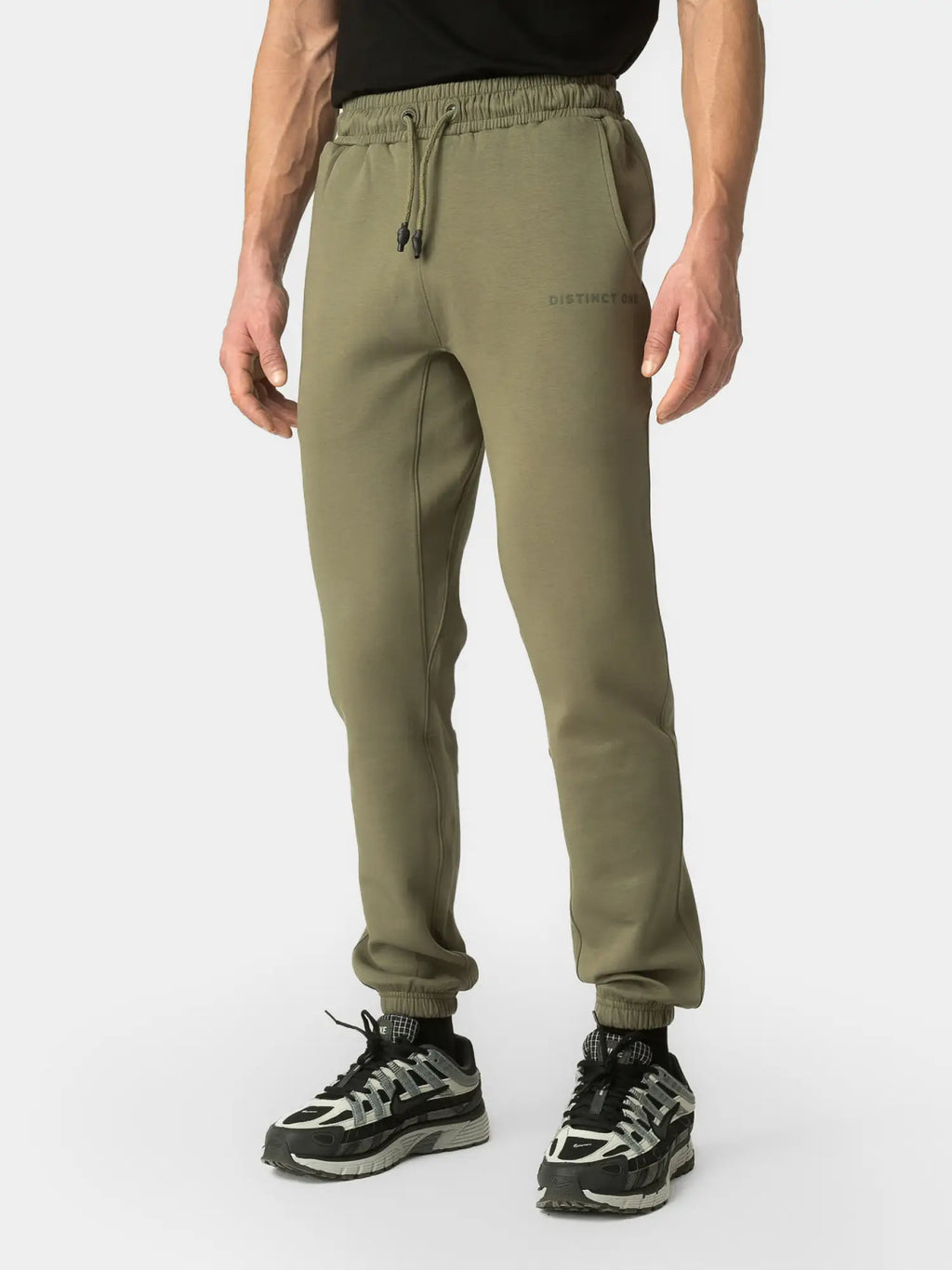 A person wearing olive green jogger sweatpants with a drawstring waist and elastic cuffs, paired with black and gray sneakers, shown against a light gray studio background.