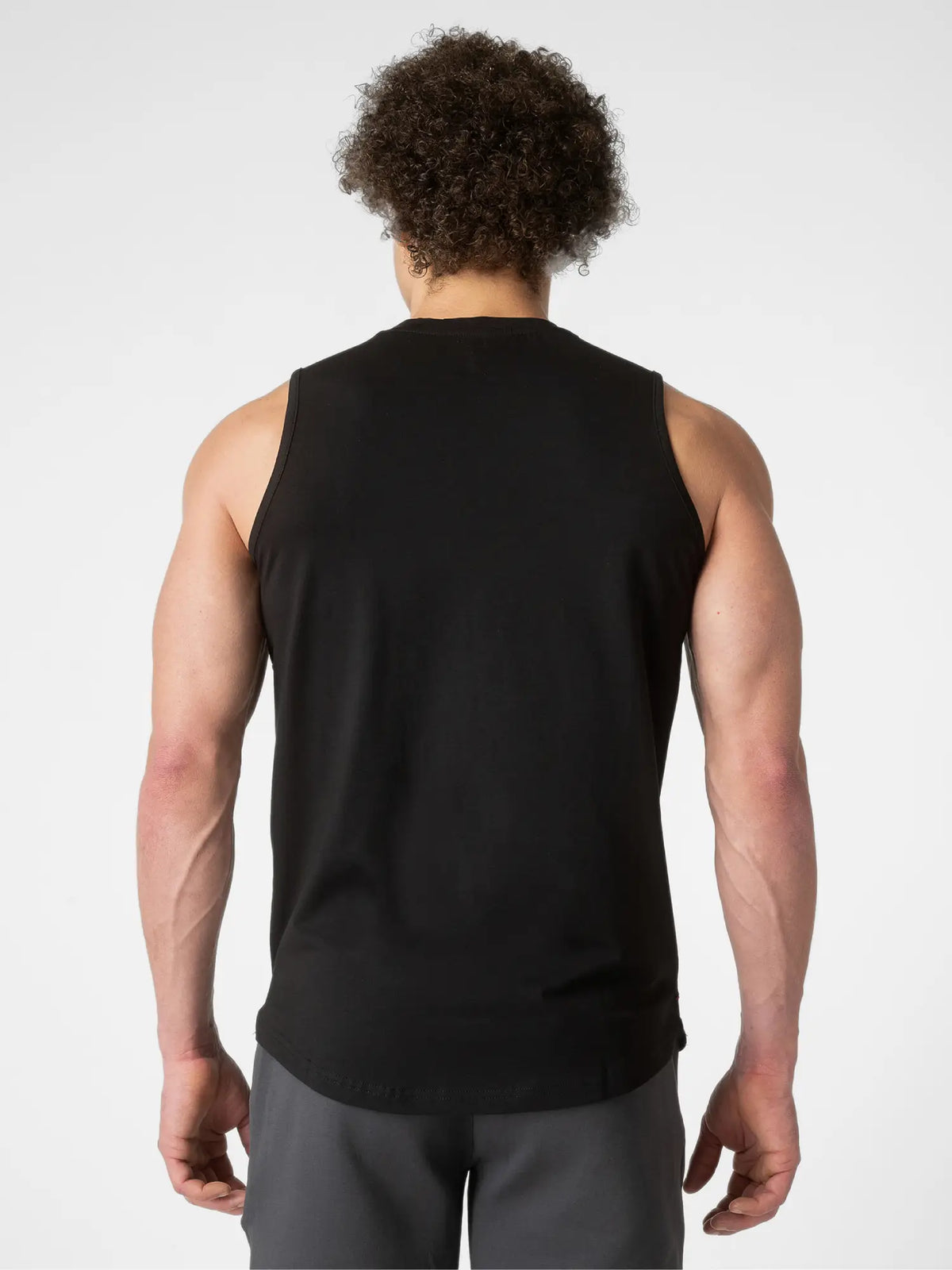 A man viewed from behind wearing a black sleeveless athletic tank top and gray shorts against a light gray studio background.