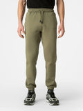 A person wearing olive green jogger sweatpants with a drawstring waist and elastic cuffs, paired with black and white sneakers, shown from the waist down on a light gray studio background.