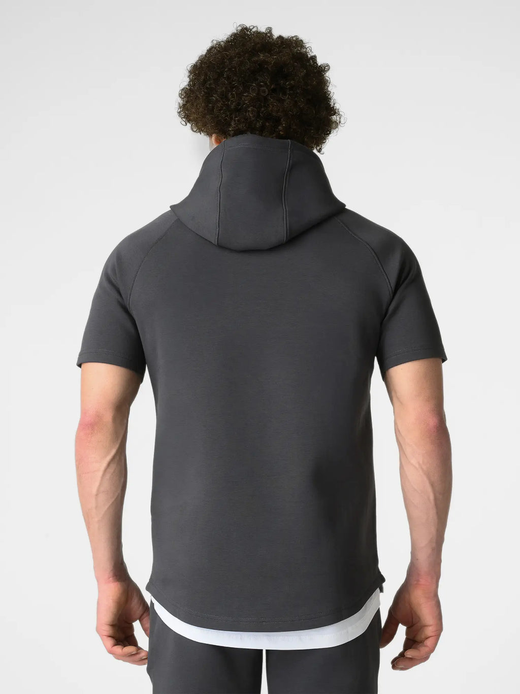 A man viewed from behind wearing a dark gray short-sleeve athletic hoodie with a high collar, paired with a white undershirt and dark gray pants, against a light gray studio background.