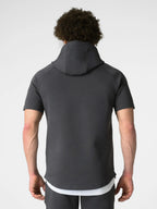 A man viewed from behind wearing a dark gray short-sleeve athletic hoodie with a high collar, paired with a white undershirt and dark gray pants, against a light gray studio background.