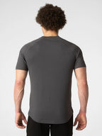 A man viewed from behind wearing a dark gray short-sleeve athletic t-shirt and black pants, standing against a light gray studio background.