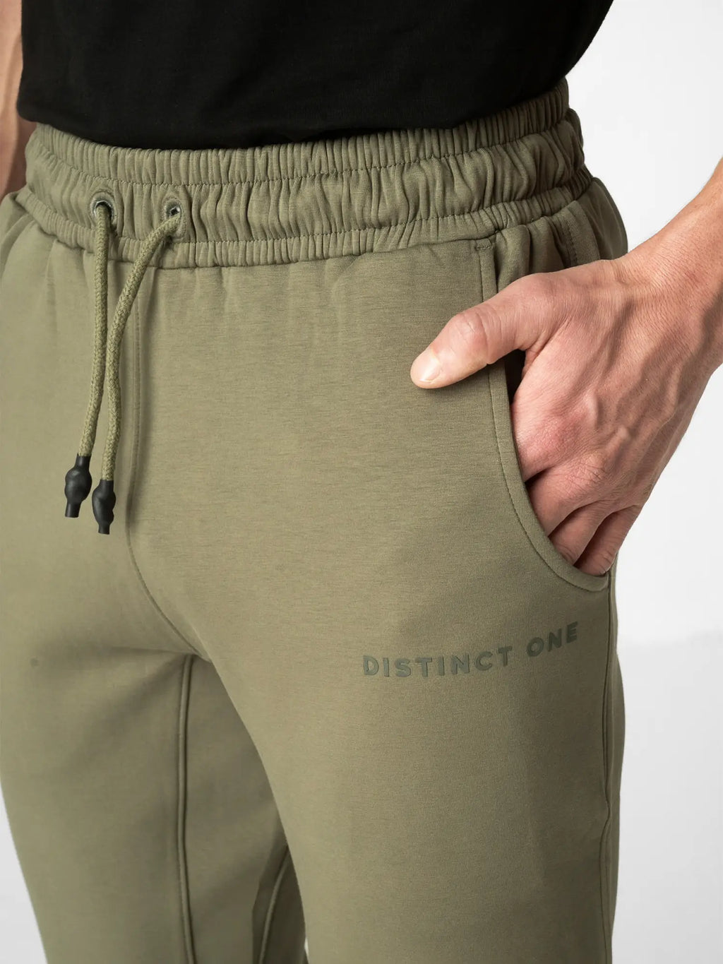 Close-up of a person wearing olive green sweatpants with an elastic drawstring waist, side pocket, and 'DISTINCT ONE' printed on the thigh, paired with a black top against a light gray background.