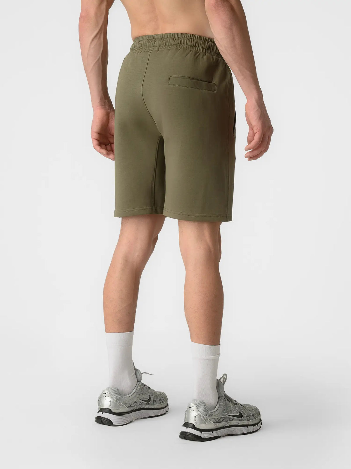 A man shown from the back wearing olive green athletic shorts, white crew socks, and silver sneakers against a plain light gray studio background.