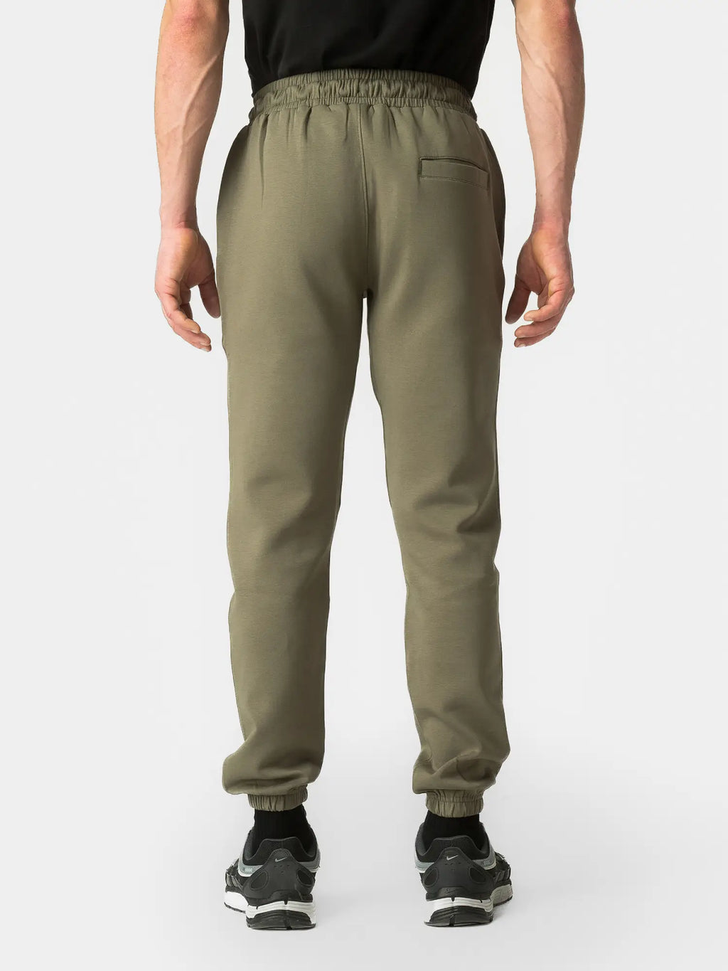 A person shown from the back wearing olive green jogger sweatpants with an elastic waistband and cuffs, paired with a black top and black sneakers, against a light gray studio background.