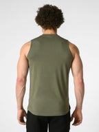A man viewed from behind wearing an olive green sleeveless tank top and black pants against a light gray studio background.