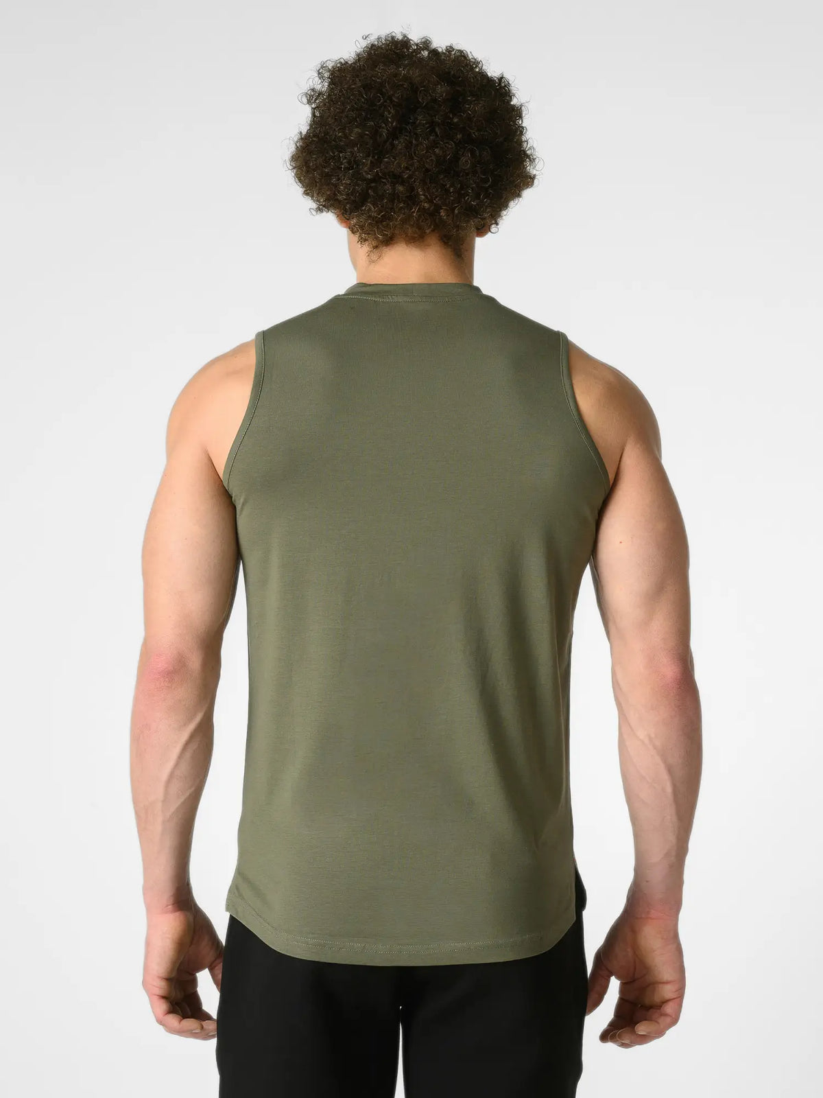 A man viewed from behind wearing an olive green sleeveless tank top and black pants against a light gray studio background.