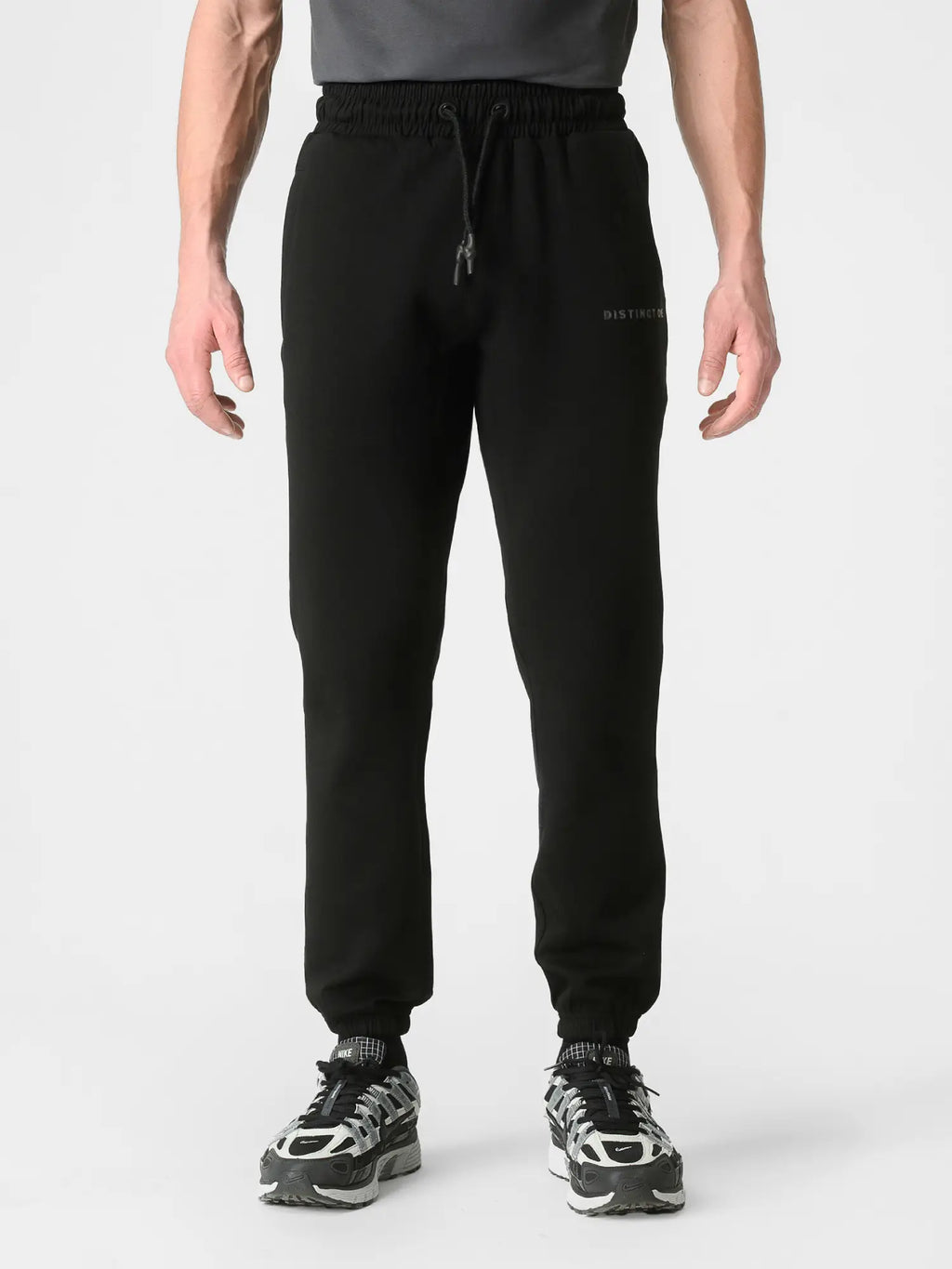 A person wearing black jogger sweatpants with a drawstring waist and elastic cuffs, paired with black and white sneakers, shown against a light gray studio background.