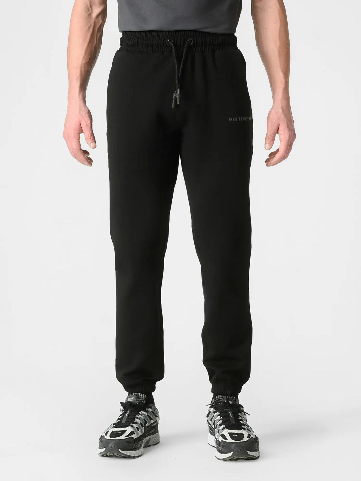 A person wearing black jogger sweatpants with a drawstring waist and elastic cuffs, paired with black and white sneakers, shown against a light gray studio background.