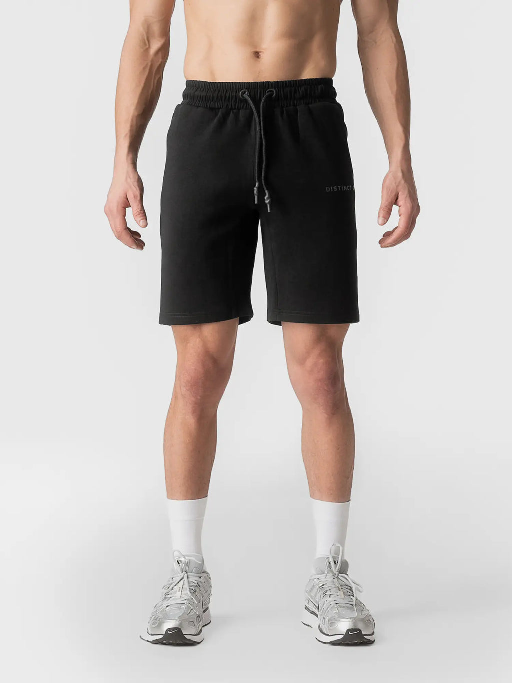 A man wearing black athletic drawstring shorts, white socks, and silver running shoes, shown from the waist down against a light gray studio background.