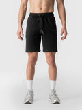 A man wearing black athletic drawstring shorts, white socks, and silver running shoes, shown from the waist down against a light gray studio background.