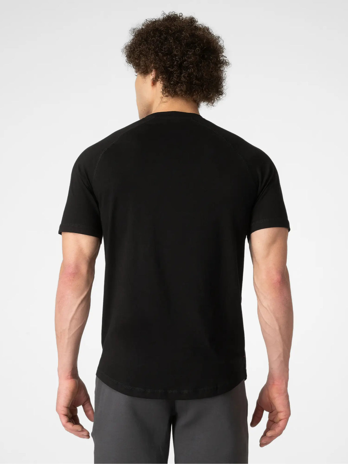A man viewed from behind wearing a black short-sleeve athletic t-shirt and gray athletic pants against a light gray studio background.