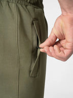 Close-up of olive green pants with a hand unzipping a side pocket, showing the fabric texture and zipper detail against a light gray background.