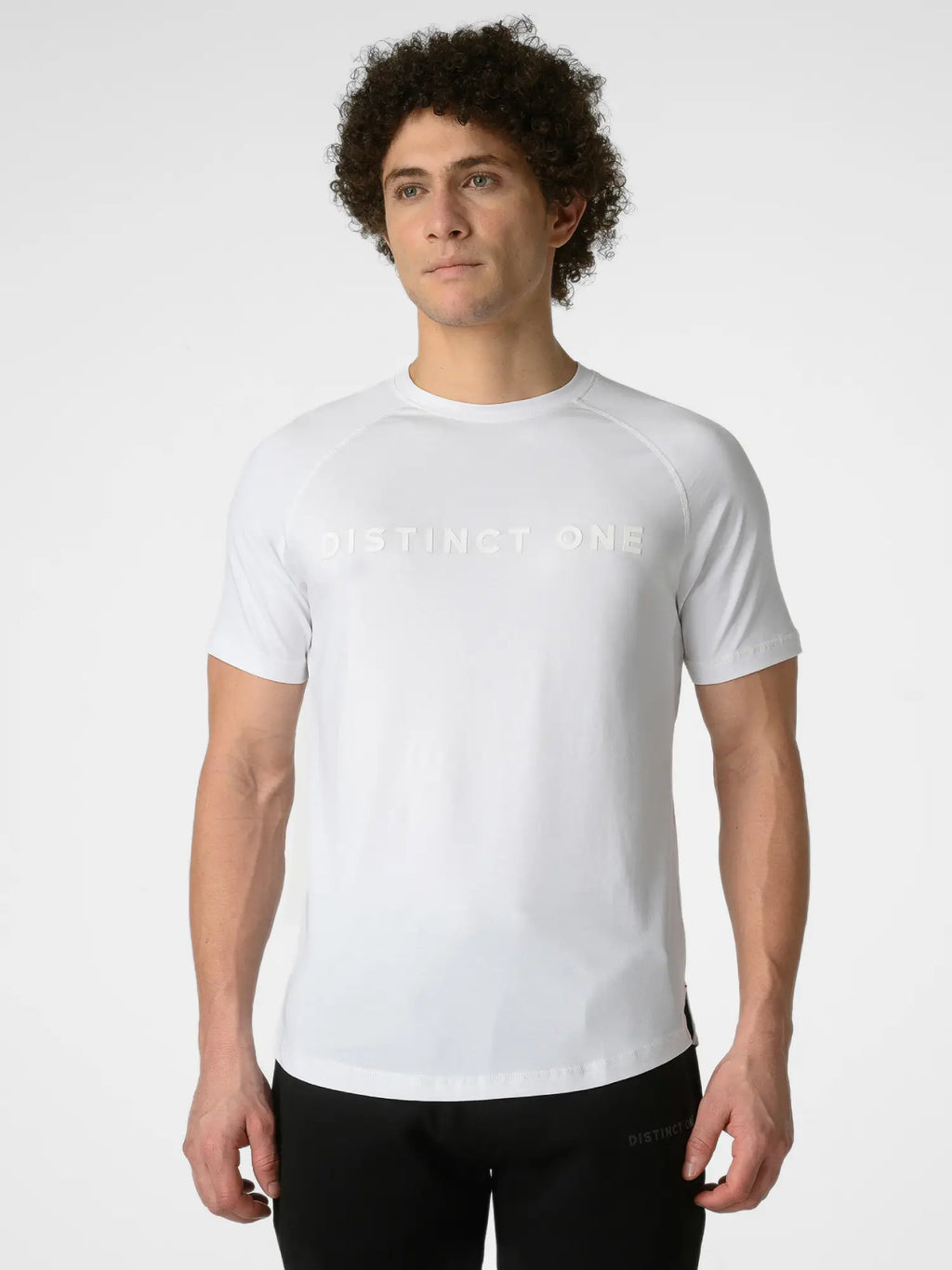 A man wearing a white short-sleeve athletic t-shirt with 'DISTINCT ONE' printed across the chest in a subtle tonal font, paired with black athletic pants, against a light gray studio background.