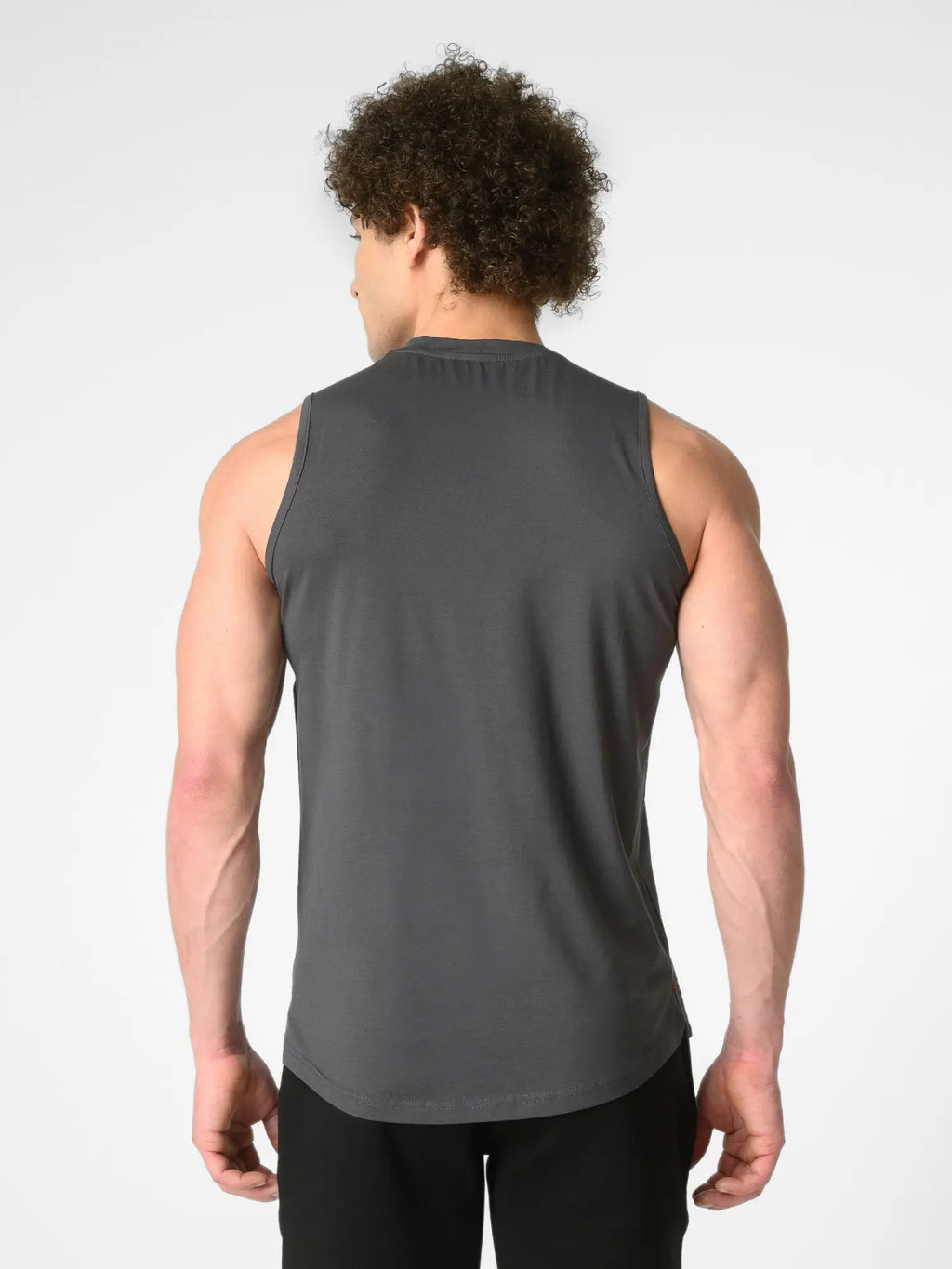 A man viewed from behind wearing a dark gray sleeveless athletic tank top and black pants, standing against a light gray studio background.