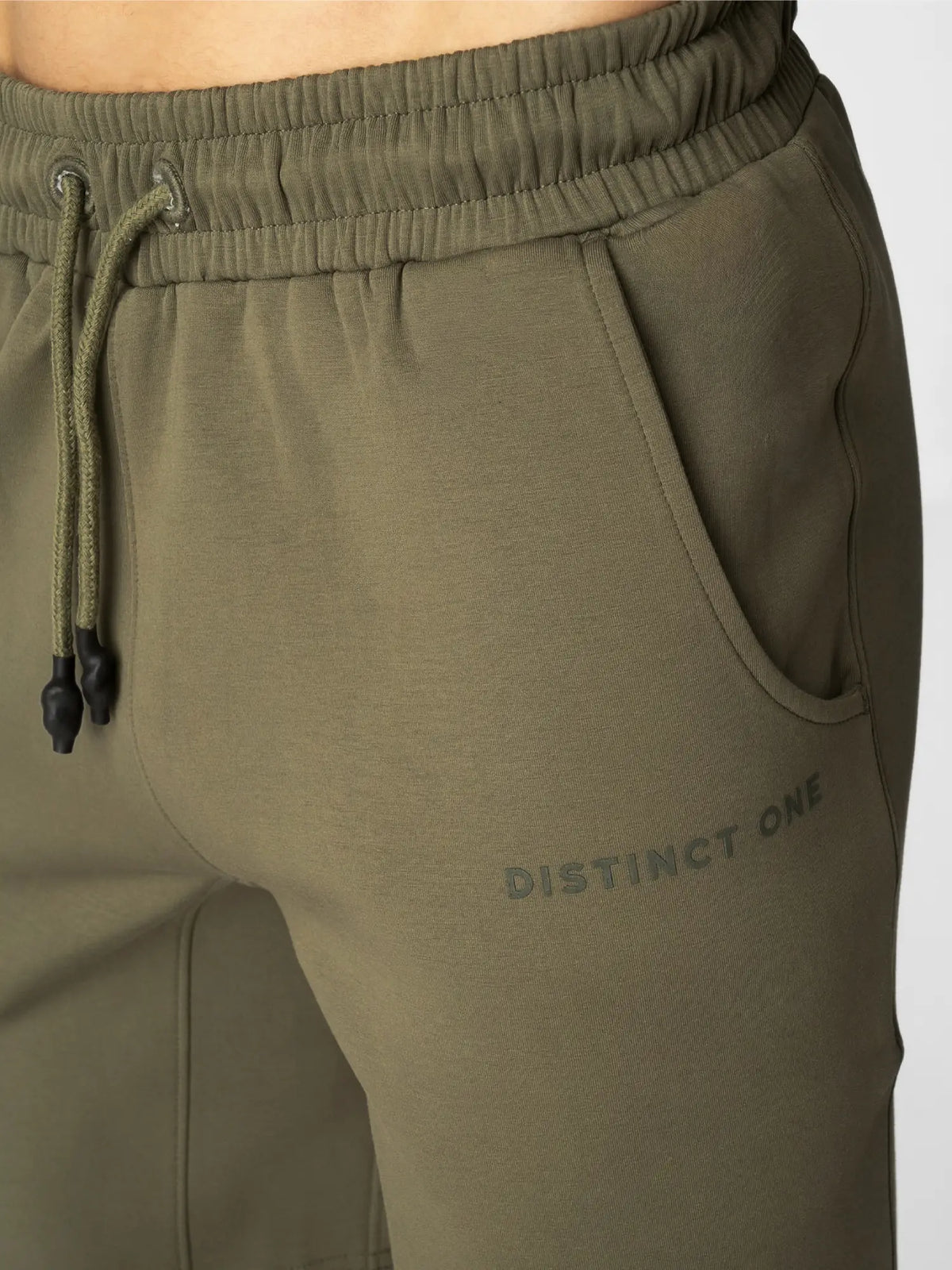 Close-up of olive green sweatpants with a drawstring waist, side pocket, and 'DISTINCT ONE' printed in dark gray on the thigh.