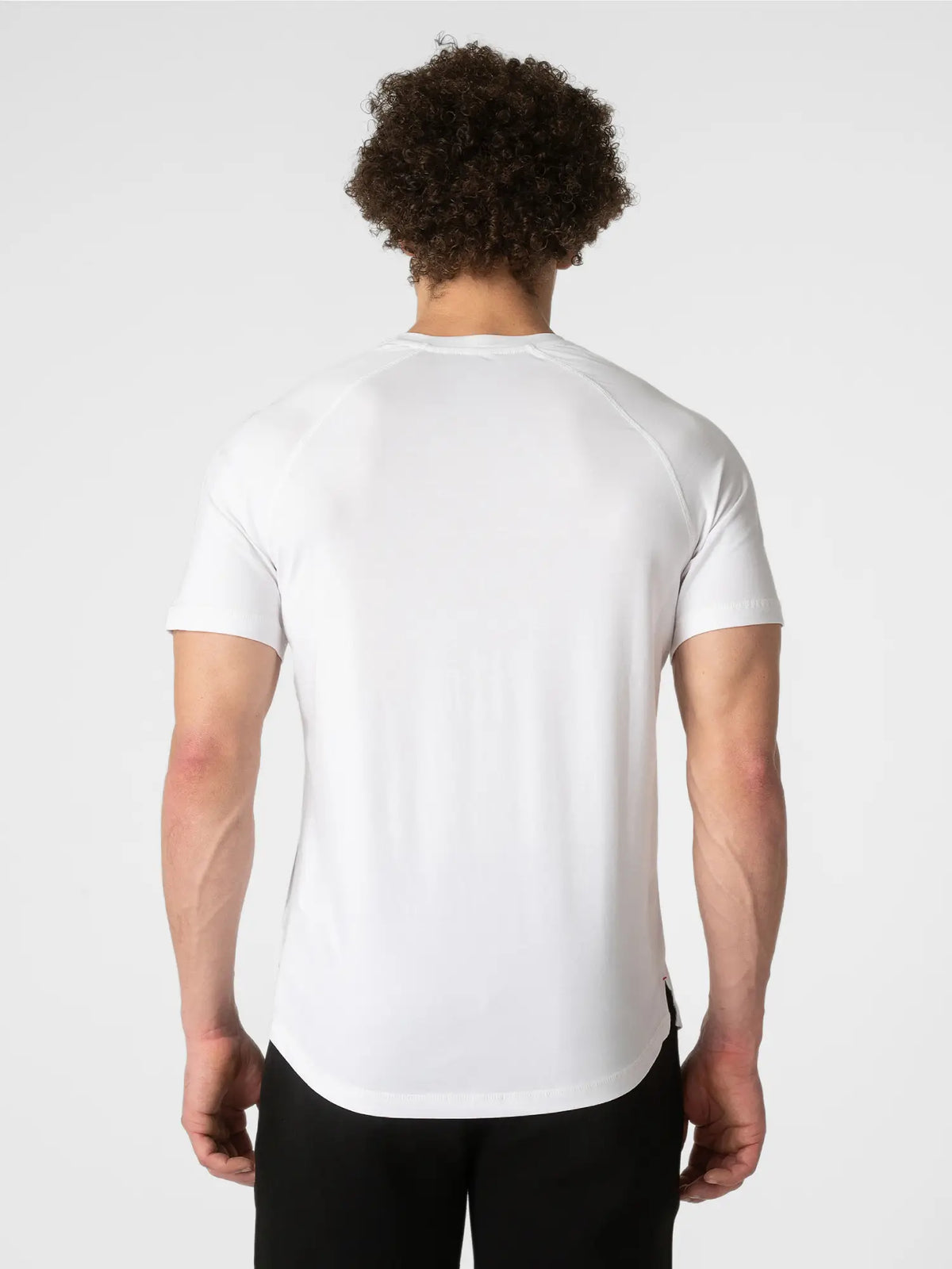 A man viewed from behind wearing a white short-sleeve athletic t-shirt and black pants, set against a plain light gray studio background.