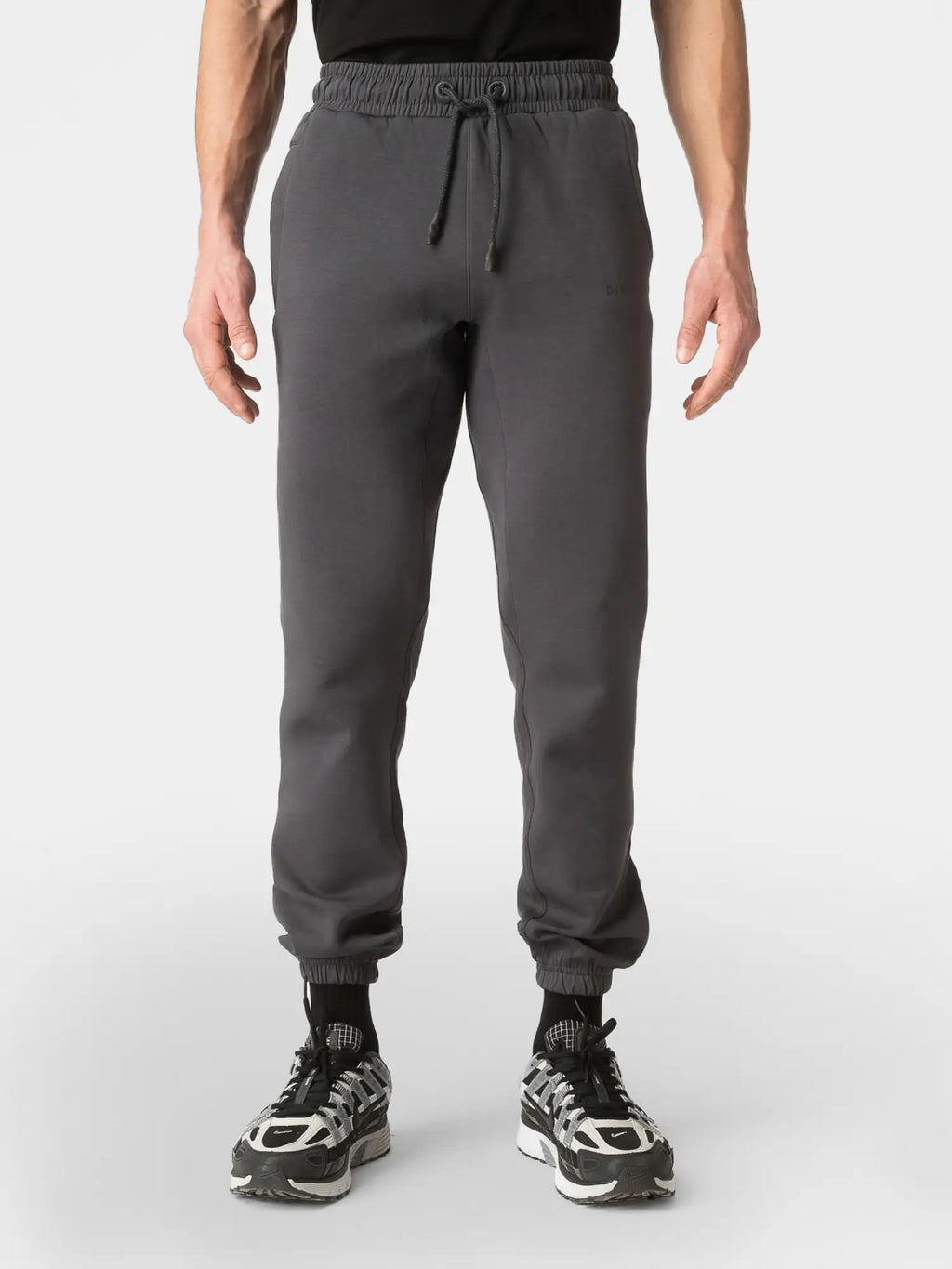 A person wearing dark gray jogger sweatpants with an elastic drawstring waist and ribbed cuffs, paired with black socks and black-and-white sneakers, shown against a light gray studio background.