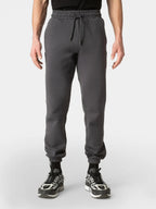 A person wearing dark gray jogger sweatpants with an elastic drawstring waist and ribbed cuffs, paired with black socks and black-and-white sneakers, shown against a light gray studio background.