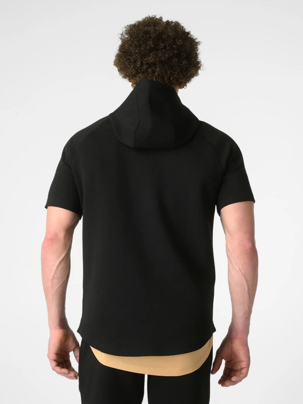 A man viewed from behind wearing a black short-sleeve hooded top with a high neck, layered over a beige undershirt. The photo is shot against a clean, light gray studio background.