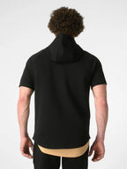 A man viewed from behind wearing a black short-sleeve hooded top with a high neck, layered over a beige undershirt. The photo is shot against a clean, light gray studio background.