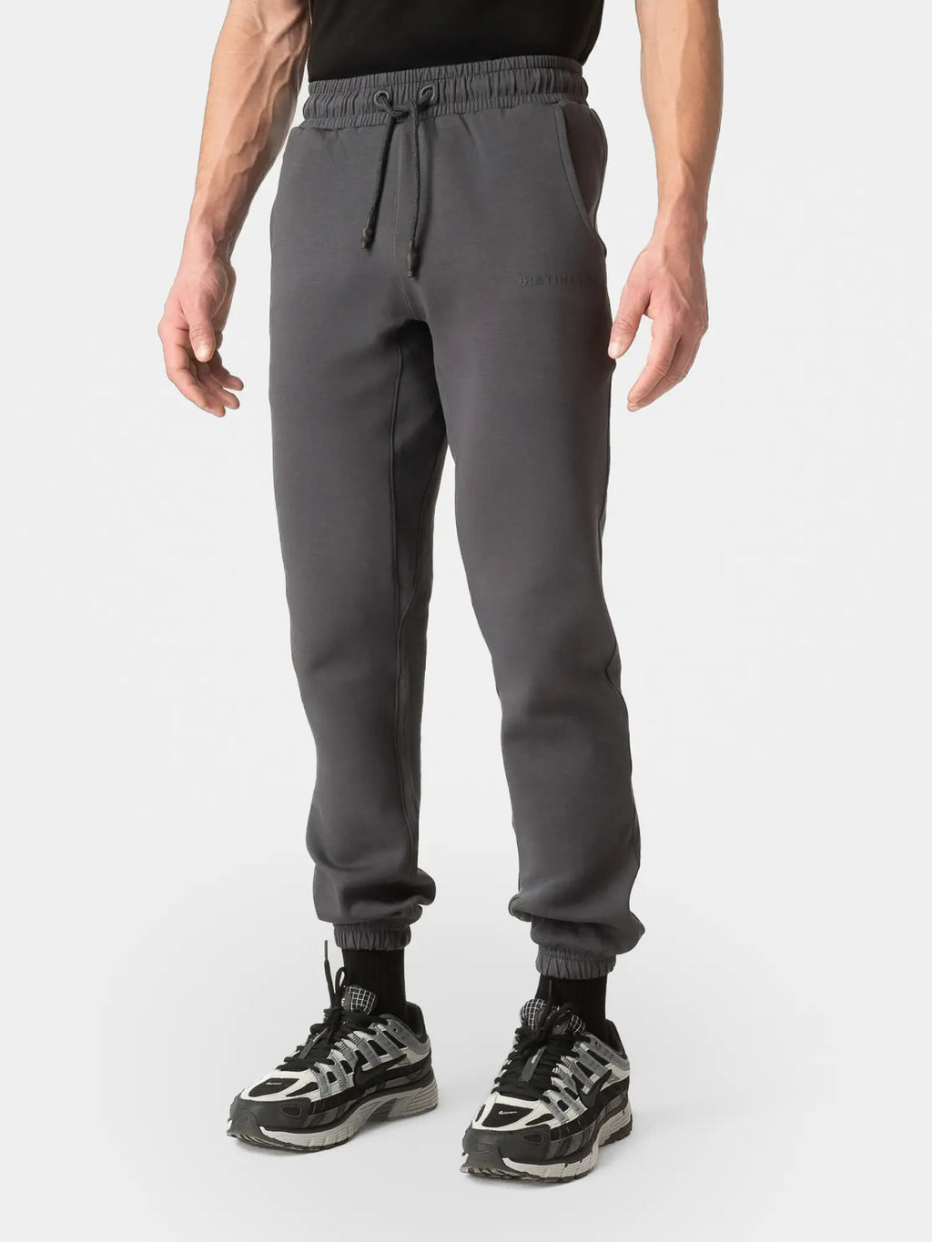 A person wearing dark gray jogger sweatpants with an elastic waistband and drawstring, paired with black and gray sneakers, shown from the waist down on a light gray studio background.