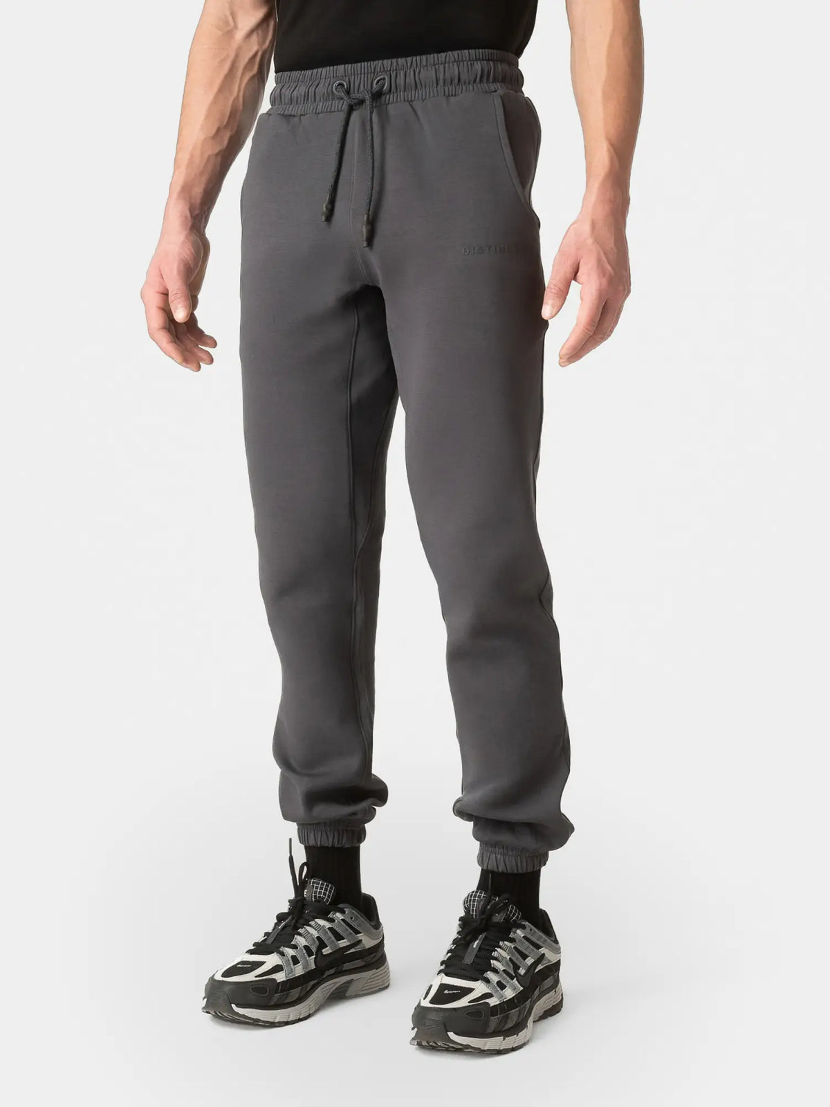 A person wearing dark gray jogger sweatpants with an elastic waistband and drawstring, paired with black and gray sneakers, shown from the waist down on a light gray studio background.