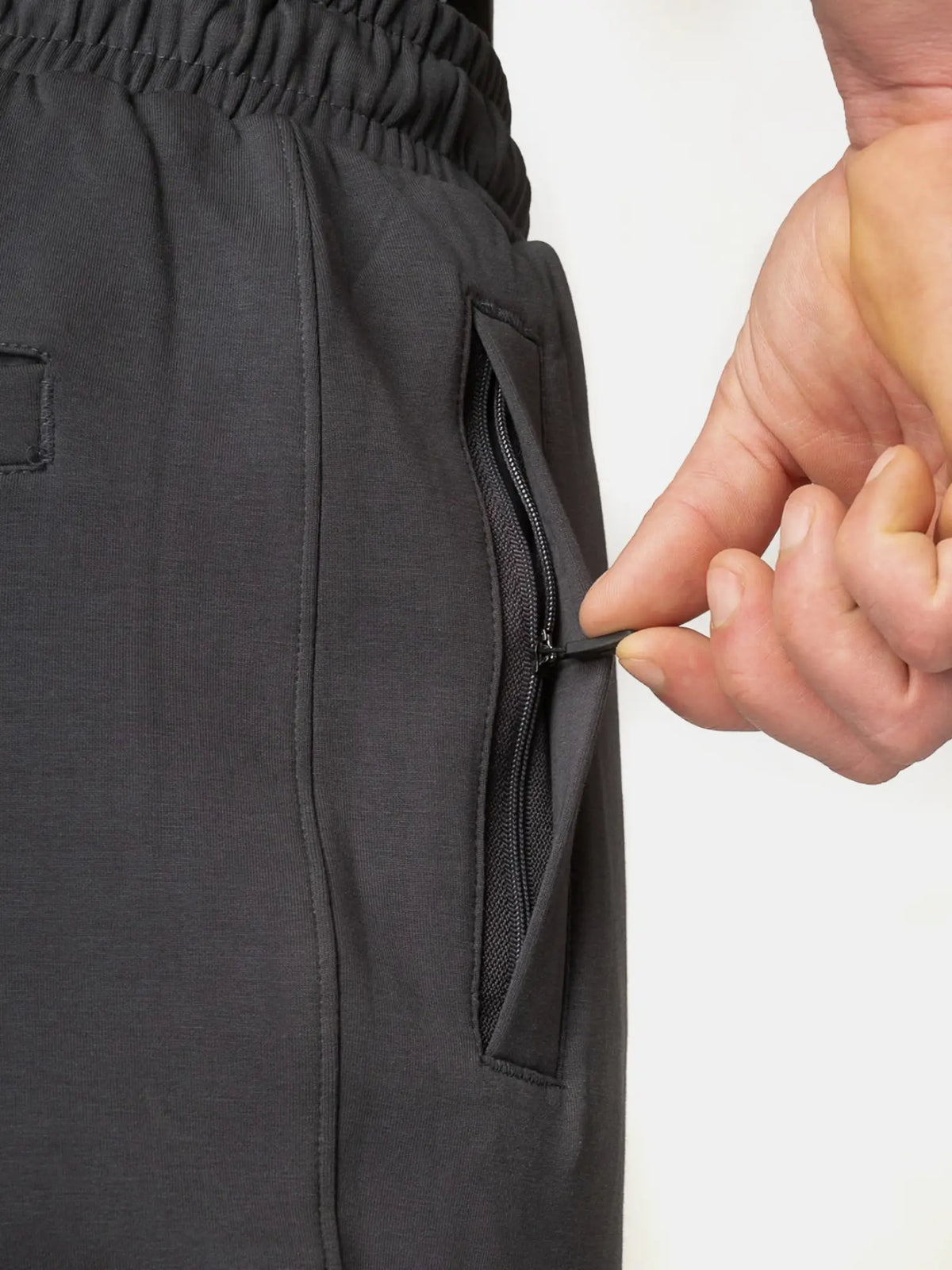 Close-up of a person's hand unzipping a side pocket of dark gray heathered athletic pants, showing the texture and zipper detail against a plain white background.