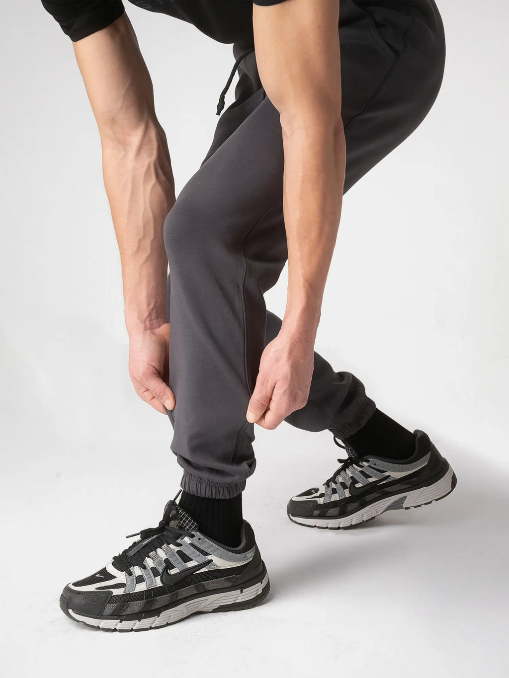 A person wearing dark gray jogger pants, black socks, and black and gray athletic sneakers, shown from the waist down in a studio setting.