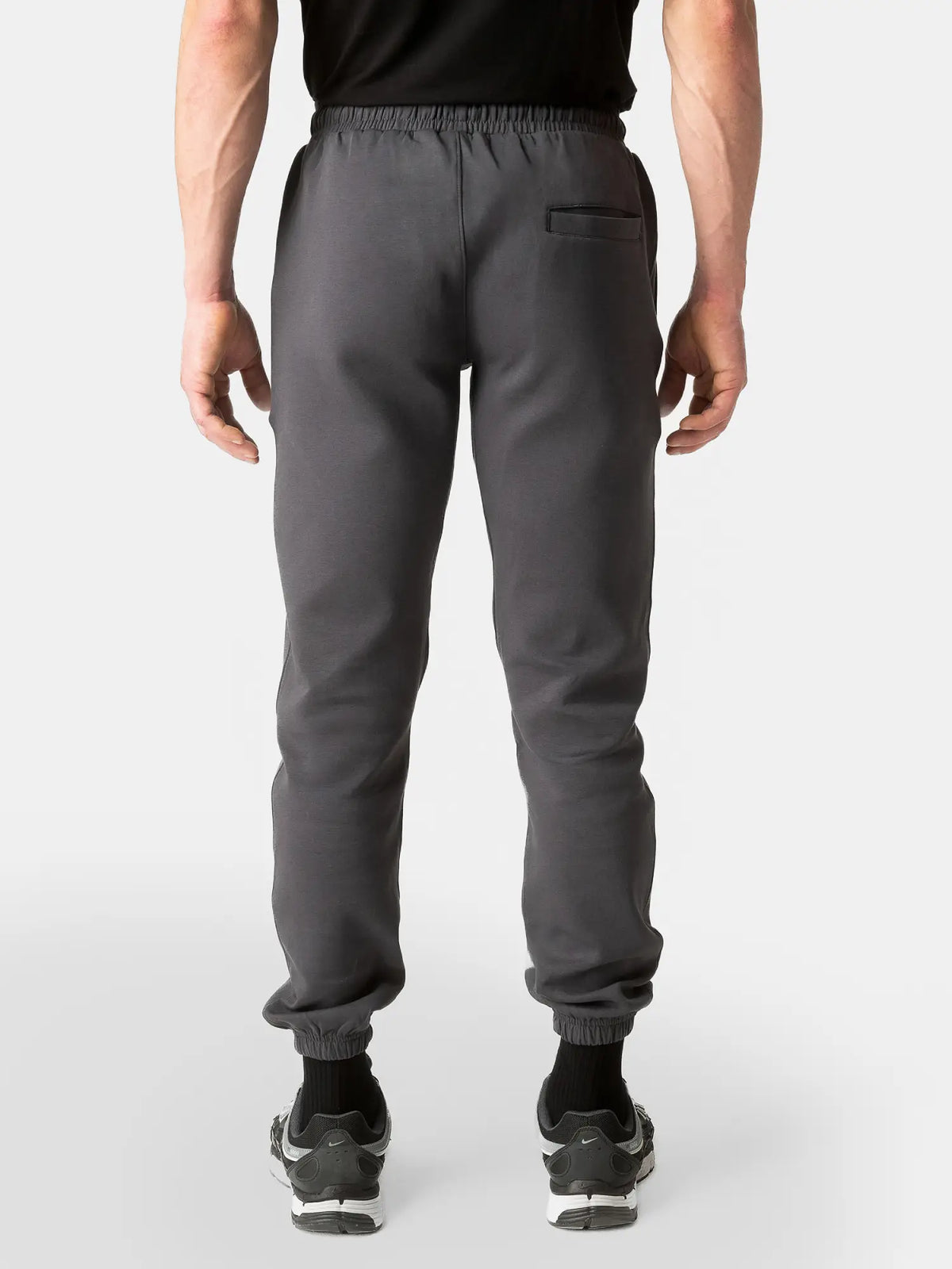 A person shown from the back wearing dark gray jogger-style pants with an elastic waistband, elastic cuffs, a side pocket, and black sneakers against a light gray studio background.