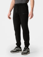 A person wearing black jogger sweatpants with a drawstring waist and a small 'DISTRICT' logo on the left thigh, paired with black and gray sneakers, against a light gray studio background.