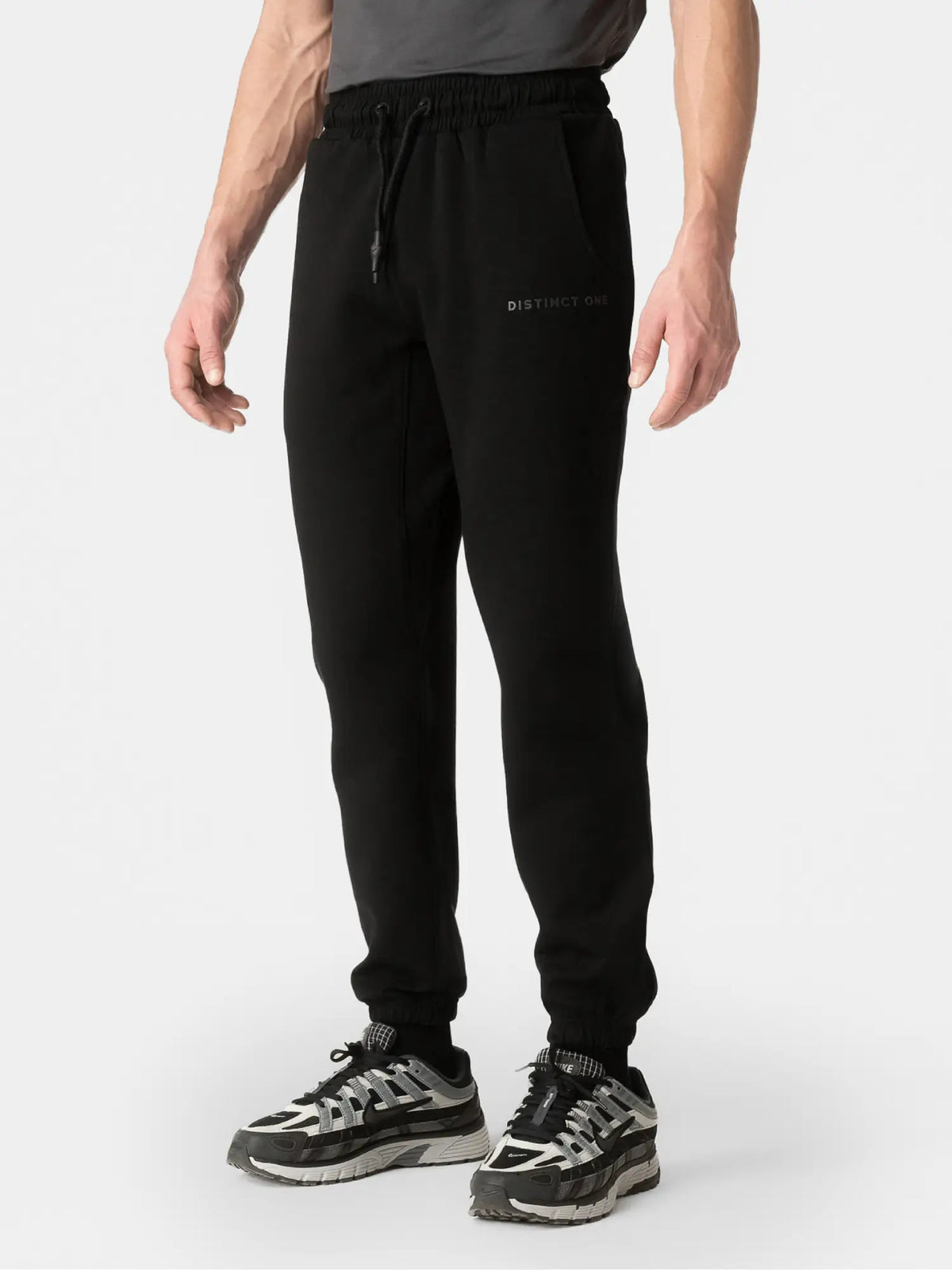 A person wearing black jogger sweatpants with a drawstring waist and a small 'DISTRICT' logo on the left thigh, paired with black and gray sneakers, against a light gray studio background.