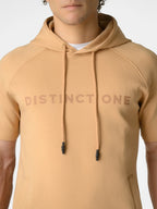 A close-up of a person wearing a tan short-sleeve hoodie with 'DISTINCTONE' printed across the chest in a darker tan tone. The hoodie has matching drawstrings with black tips and a white undershirt visible at the collar.