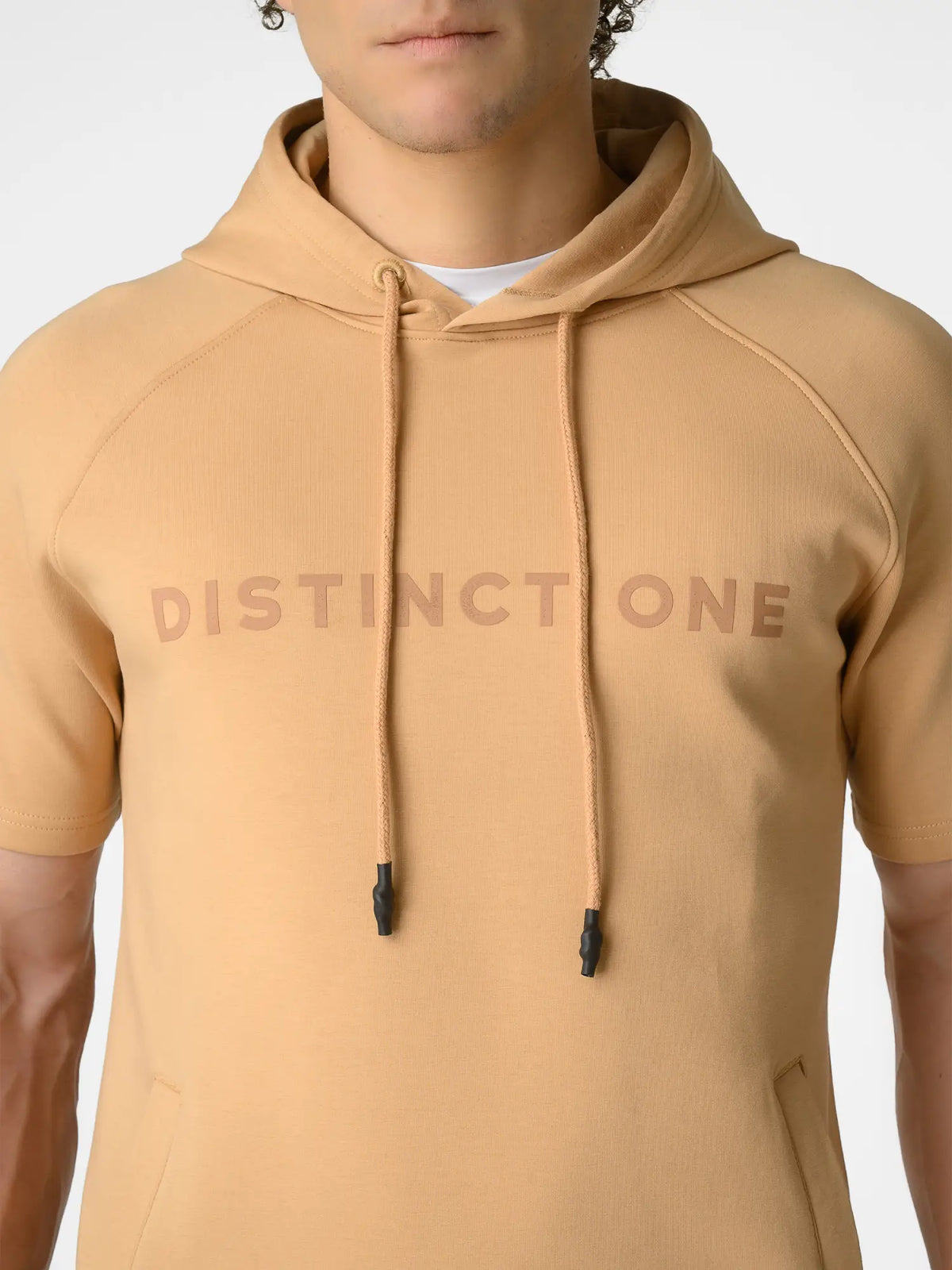 A close-up of a person wearing a tan short-sleeve hoodie with 'DISTINCTONE' printed across the chest in a darker tan tone. The hoodie has matching drawstrings with black tips and a white undershirt visible at the collar.
