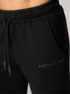 Close-up of black sweatpants with an elastic drawstring waistband and side pocket, featuring the text 'DISTINCT ONE' printed in tonal black on the lower leg.