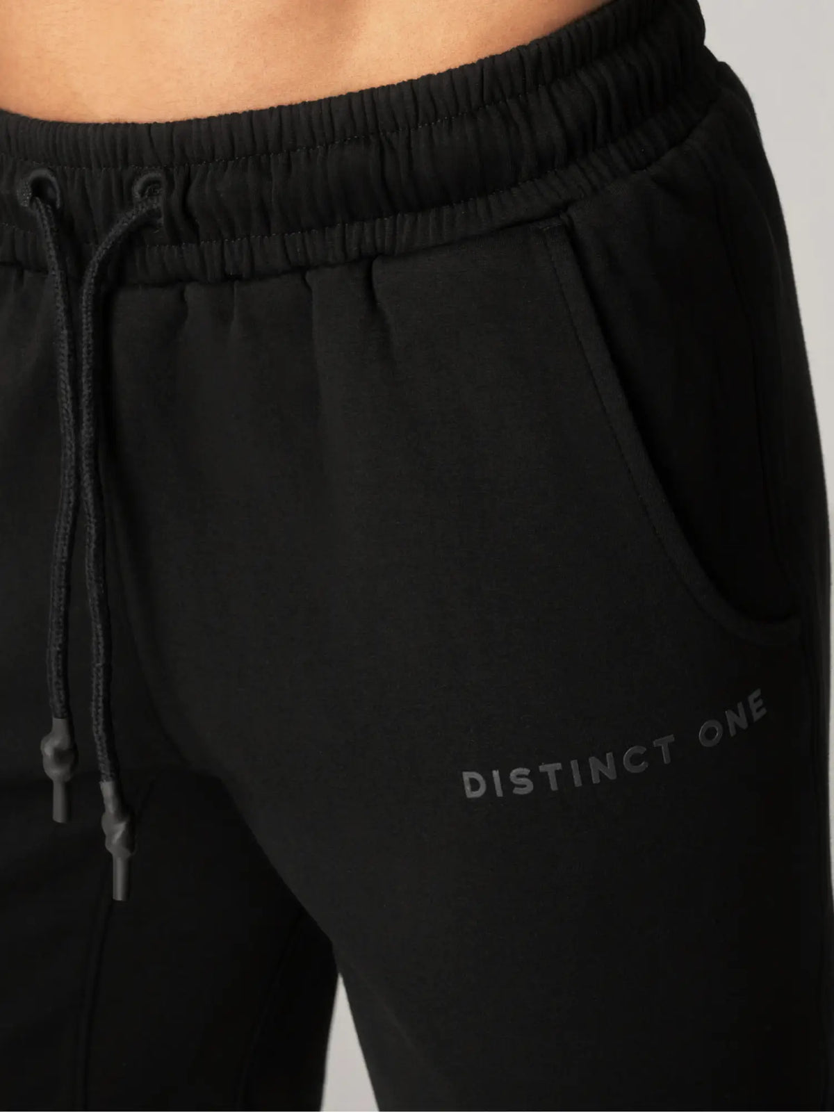 Close-up of black sweatpants with an elastic drawstring waistband and side pocket, featuring the text 'DISTINCT ONE' printed in tonal black on the lower leg.