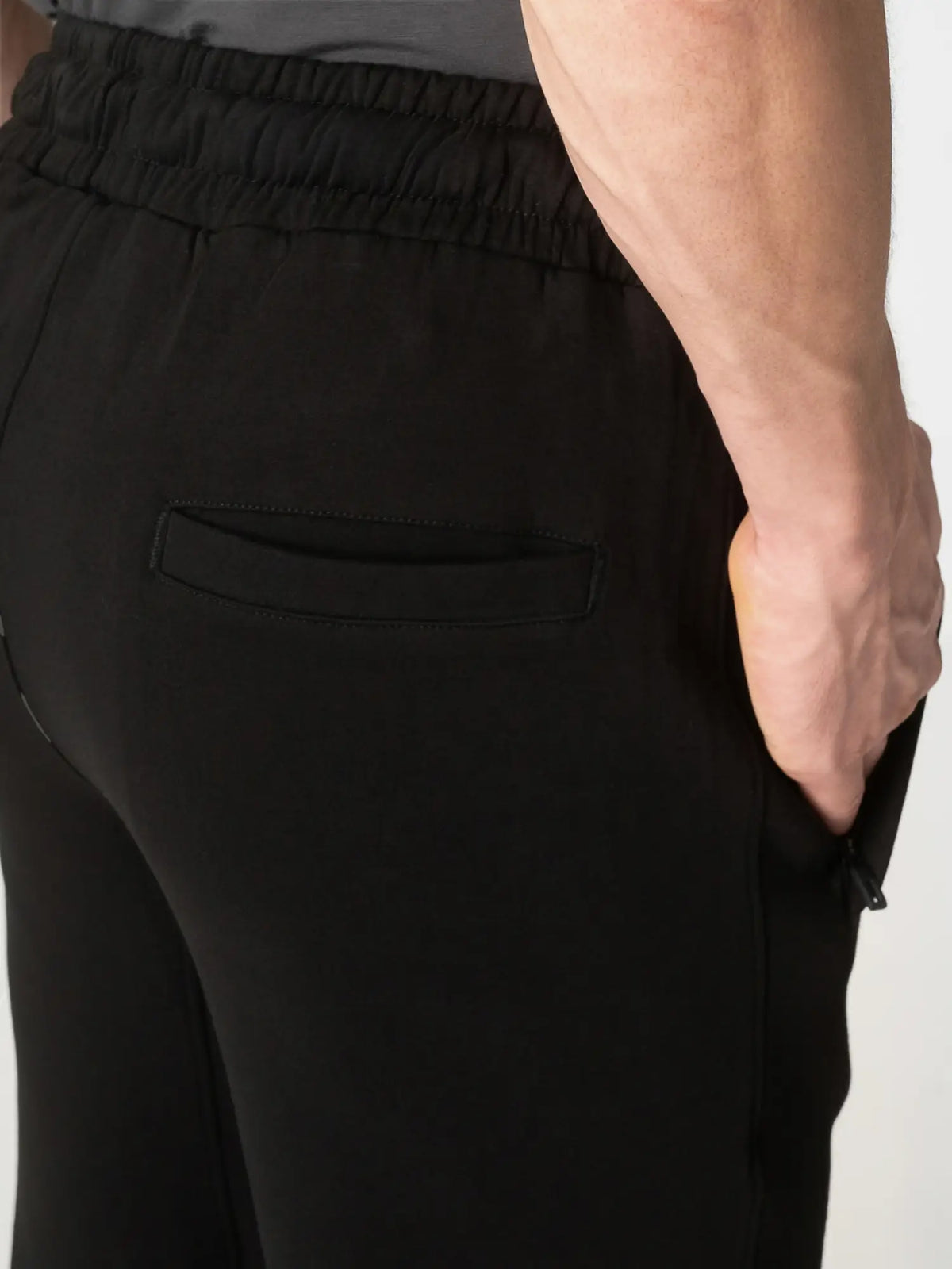 Close-up of a person wearing black elastic-waist pants, showing the side pocket and hand in the pocket against a plain light background.